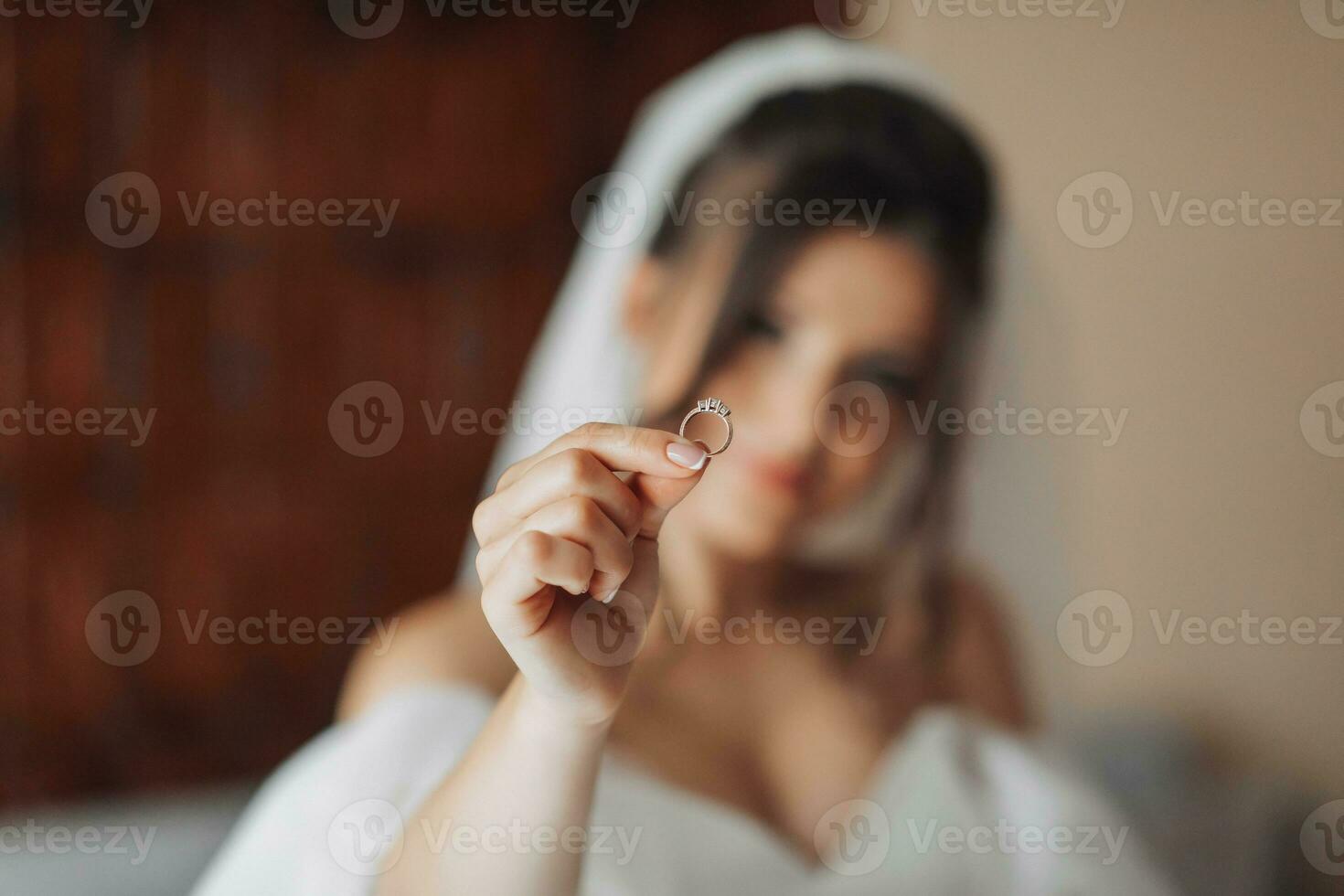 un' brunetta sposa nel un' bianca vestito detiene sua argento nozze squillo. sfocato foto, messa a fuoco su nozze squillo. francese manicure. Aperto le spalle. bellissimo mani. lungo velo. mattina di il sposa. dettagli foto