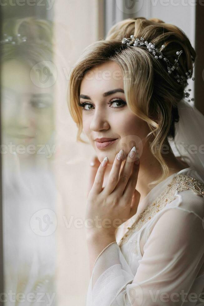 bionda sposa con elegante pettinatura e tiara, indossare un' veste, in posa, guardare a il telecamera, toccante sua viso. bellissimo trucco. elegante acconciatura. francese manicure foto