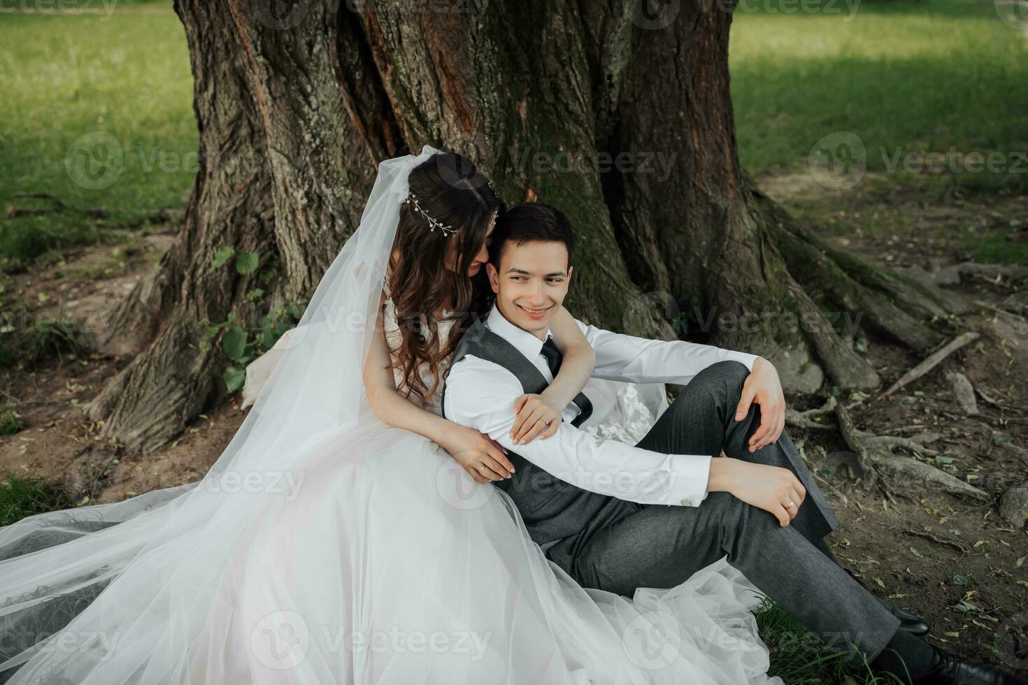 un' bellissimo sposa con lungo Riccio capelli nel un' elegante vestito abbracci il sposo, sorrisi, guardare in il lente sotto un' grande albero. ritratto di il sposa e sposo. primavera nozze. naturale trucco foto