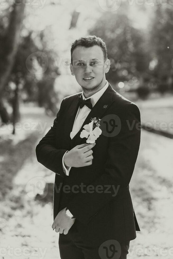 giovane elegante sposo nel il foresta con un' boutonniere nel il suo mano e un' arco legare. nero e bianca foto