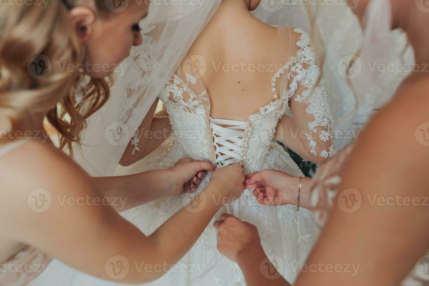 il della sposa amici allacciare il bianca voluminoso vestito di il sposa. posteriore Visualizza. Aperto le spalle. bellissimo mani. lungo velo. mattina di il sposa. foto