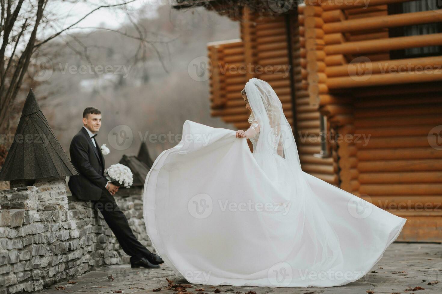 un' bellissimo e affascinante sposa è girando vicino moderno di legno case nel il parco, il sposo è Guardando. lussuoso vestito con un' lungo treno. elegante giovane sposo foto