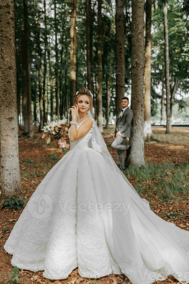 un' bellissimo giovane donna nel un' nozze vestito fra alto alberi nel il foresta con un' reale acconciatura e un' elegante tiara con un' mazzo di fiori nel sua mani, un' nozze nel d'oro colore foto