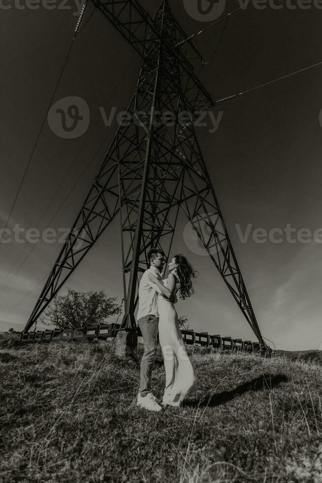 elegante modello coppia nel il montagne nel estate. un' giovane ragazzo e ragazza nel un' bianca seta vestito siamo in piedi vicino grande strutture di energia Linee. nero e bianca contrasto foto