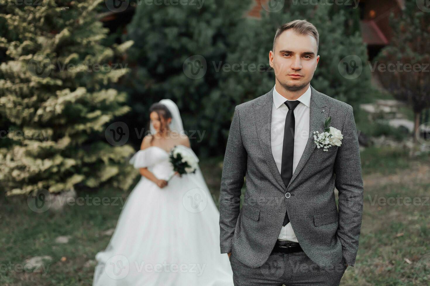 ritratto di il sposa e sposo nel natura. il sposo è nel il primo piano, il sposa è in piedi dietro a lui nel un' bianca voluminoso vestire, Tenere un' mazzo e guardare fuori uso. elegante sposo foto