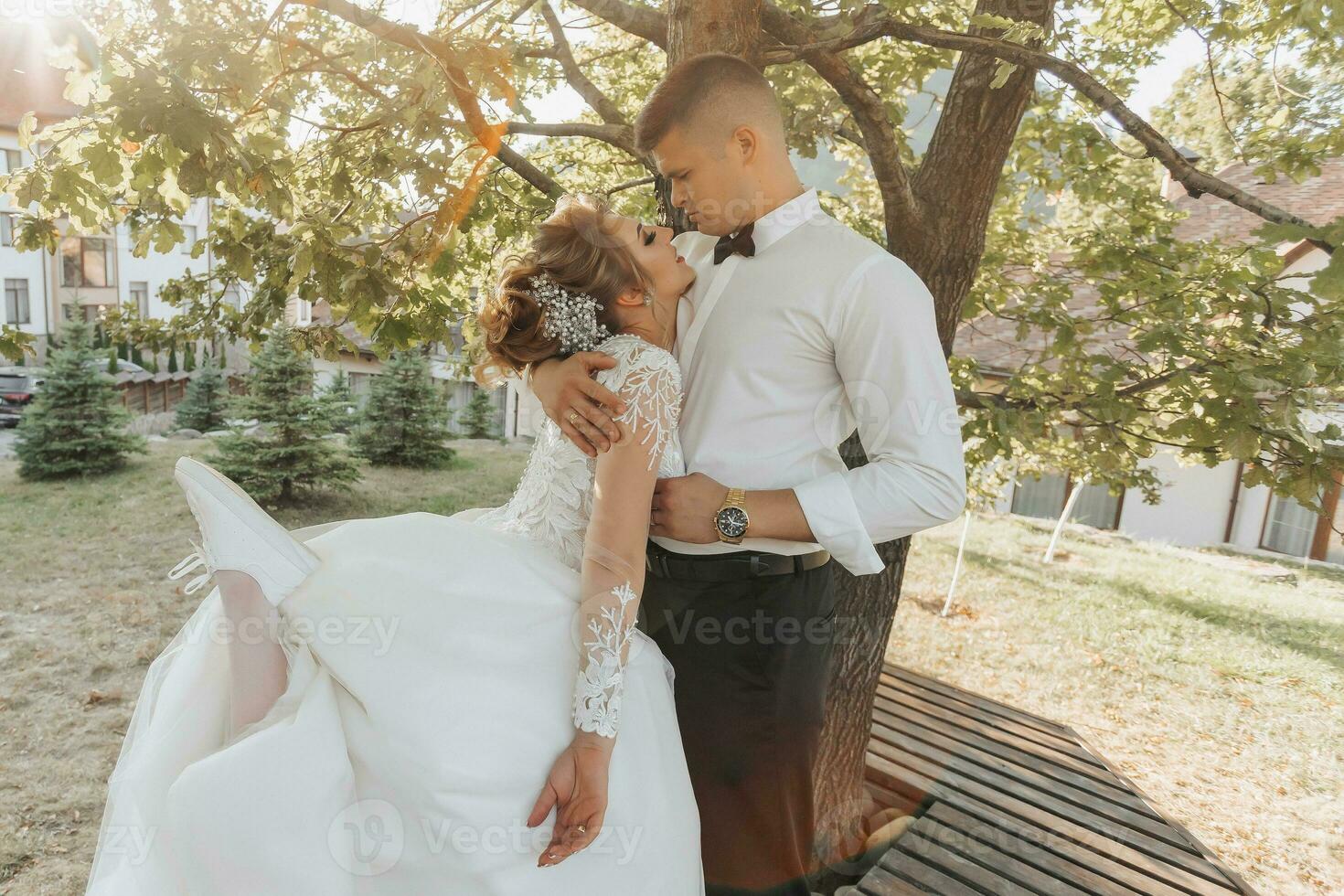 un' sposo nel un' nero completo da uomo e un' bionda sposa nel un' vestito e scarpe da ginnastica siamo in piedi, abbracciare e baci sotto un' albero. foto sessione nel natura. il foto Spettacoli un' bellissimo raggio di luce del sole. bellissimo capelli