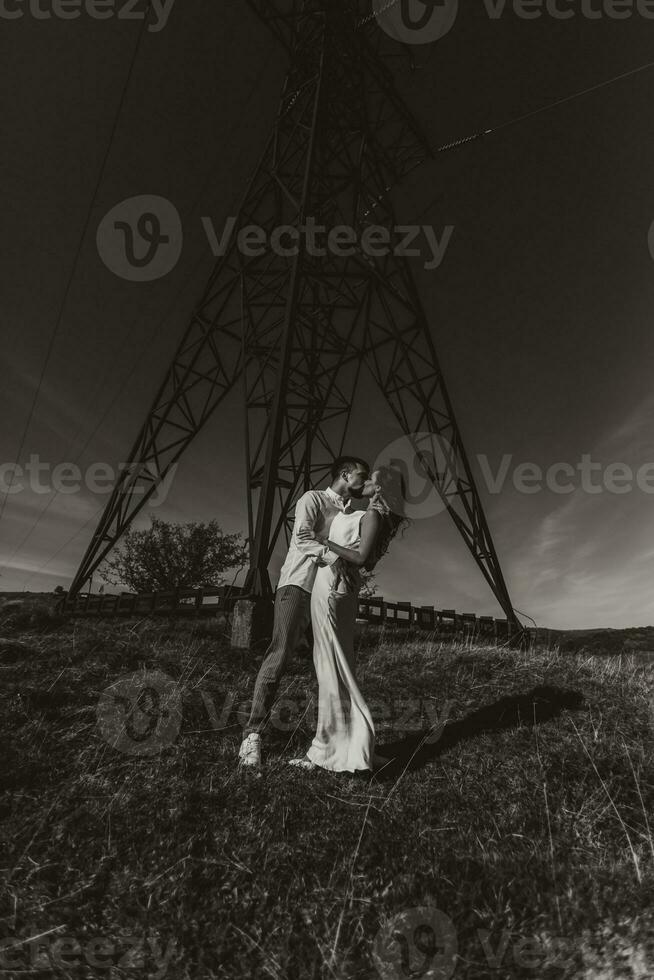 elegante modello coppia nel il montagne nel estate. un' giovane ragazzo e ragazza nel un' bianca seta vestito siamo in piedi vicino grande strutture di energia Linee. nero e bianca contrasto foto