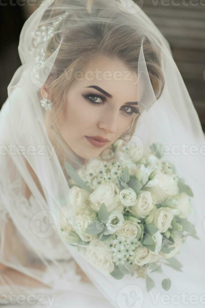 Riccio bionda sposa nel un' bianca vestire, coperto con un' velo, pose per il telecamera con un' mazzo di Rose. ritratto di il sposa. bellissimo trucco e capelli. nozze nel natura foto