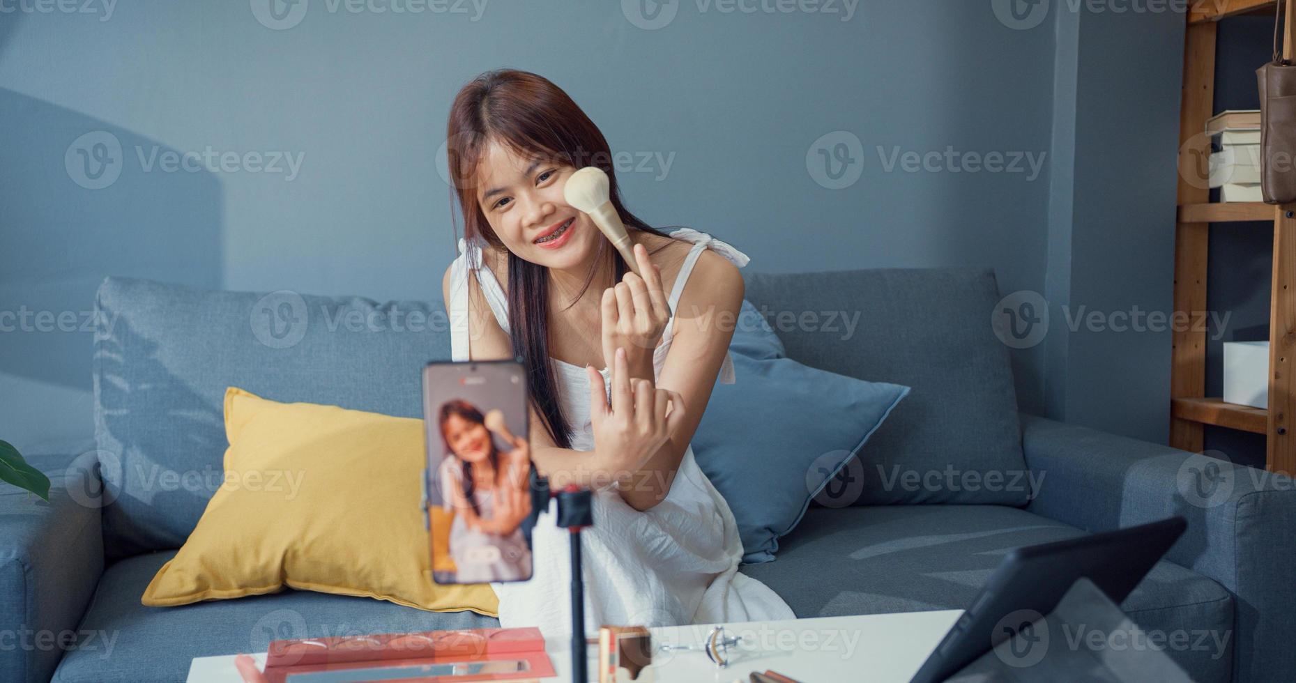 felice giovane ragazza asiatica trucco vlogger davanti alla fotocamera del telefono godere di parlare di revisione con il pubblico nel soggiorno a casa. concetto di pandemia di coronavirus a distanza sociale. libertà e concetto di stile di vita attivo. foto