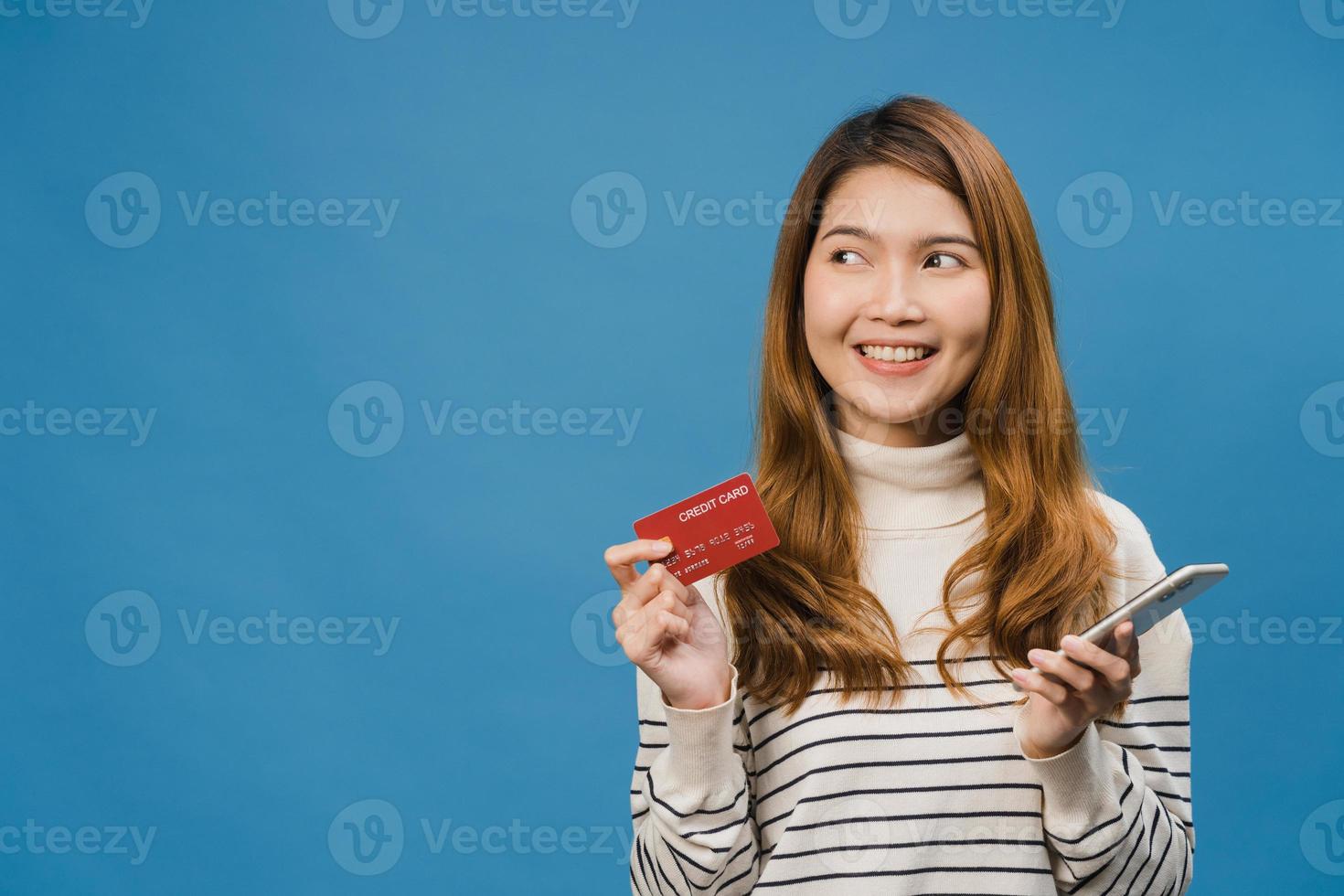 giovane donna asiatica che utilizza il telefono e la carta di credito con espressione positiva, sorride ampiamente, vestita con abiti casual e stand isolato su sfondo blu. felice adorabile donna felice esulta successo. foto