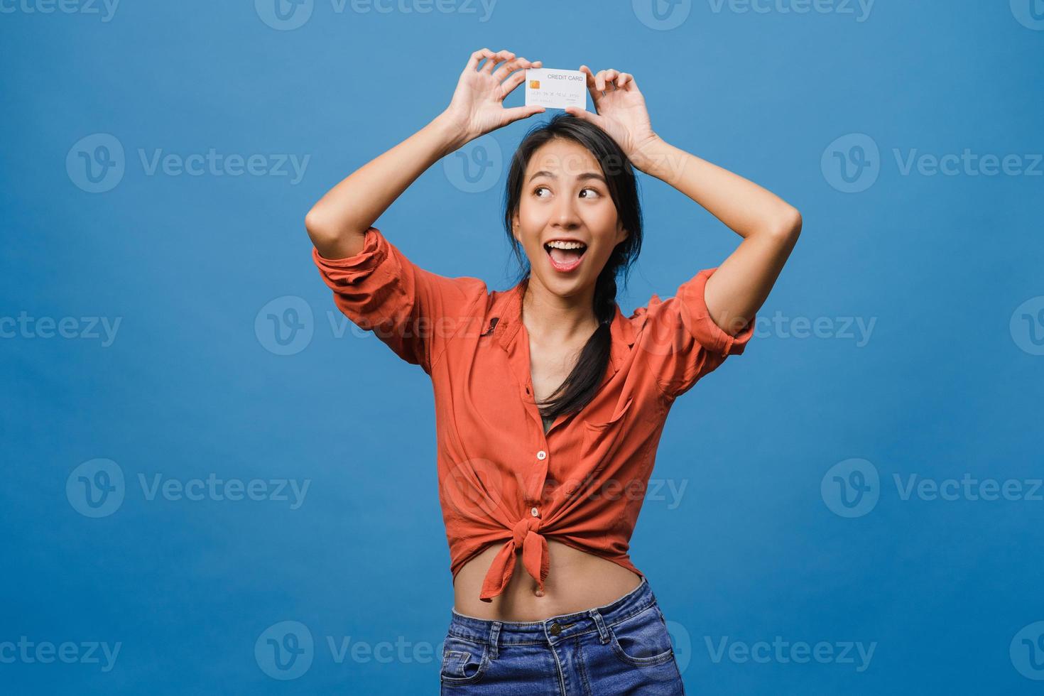 la giovane donna asiatica mostra la carta di credito con l'espressione positiva, sorride ampiamente, vestita con abiti casual sentendo felicità e stando isolata su sfondo blu. concetto di espressione facciale. foto