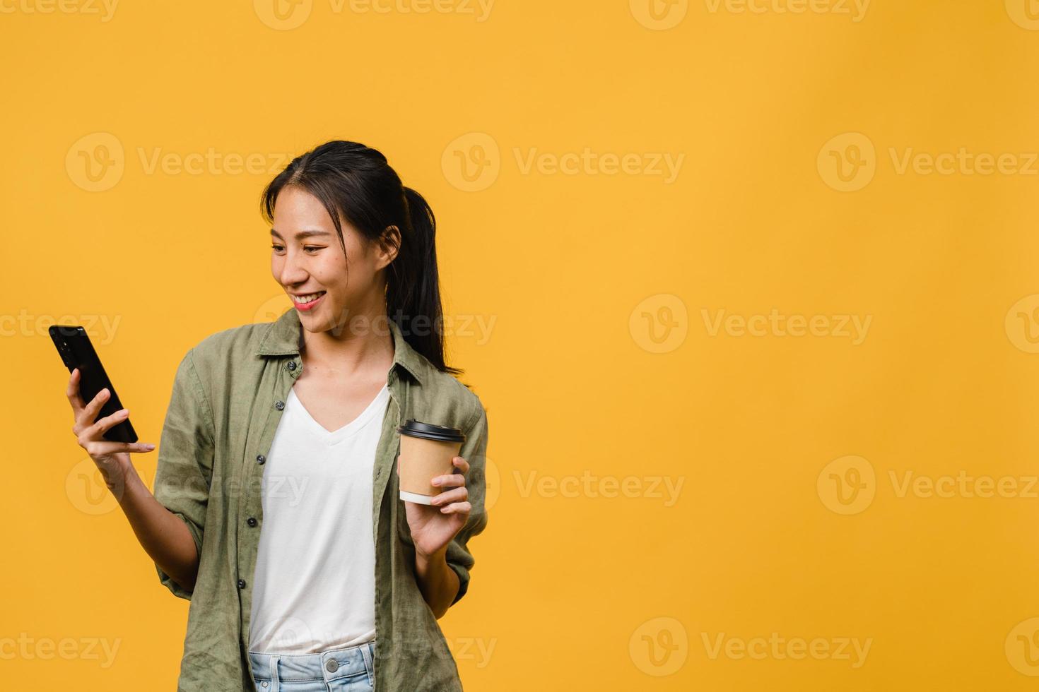 giovane donna asiatica che usa il telefono e tiene la tazza di caffè con espressione positiva, sorride ampiamente, vestita con un panno casual sentendo felicità e stando isolato su sfondo giallo. concetto di espressione facciale. foto