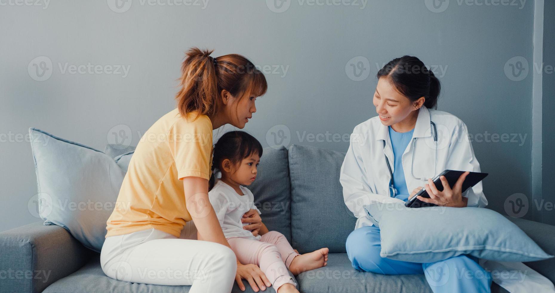 giovane donna pediatra asiatica medico e bambina paziente utilizzando tablet digitale condividendo buone notizie di test di salute con mamma felice sedersi sul divano in casa. assicurazione medica, visita il concetto di paziente a casa. foto