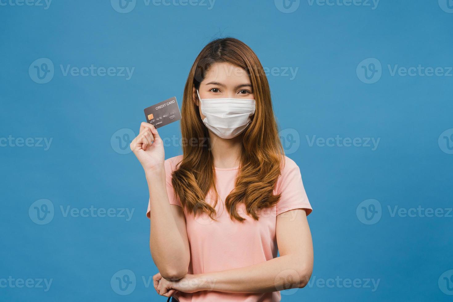 la giovane donna asiatica che indossa la maschera medica mostra la carta di credito con espressione positiva, sorride ampiamente, vestita con abiti casual sentendosi felice e stando isolata su sfondo blu. foto
