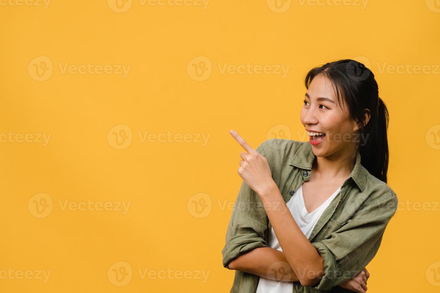 ritratto di giovane donna asiatica sorridente con espressione allegra, mostra qualcosa di straordinario in uno spazio vuoto in abbigliamento casual e in piedi isolato su sfondo giallo. concetto di espressione facciale. foto