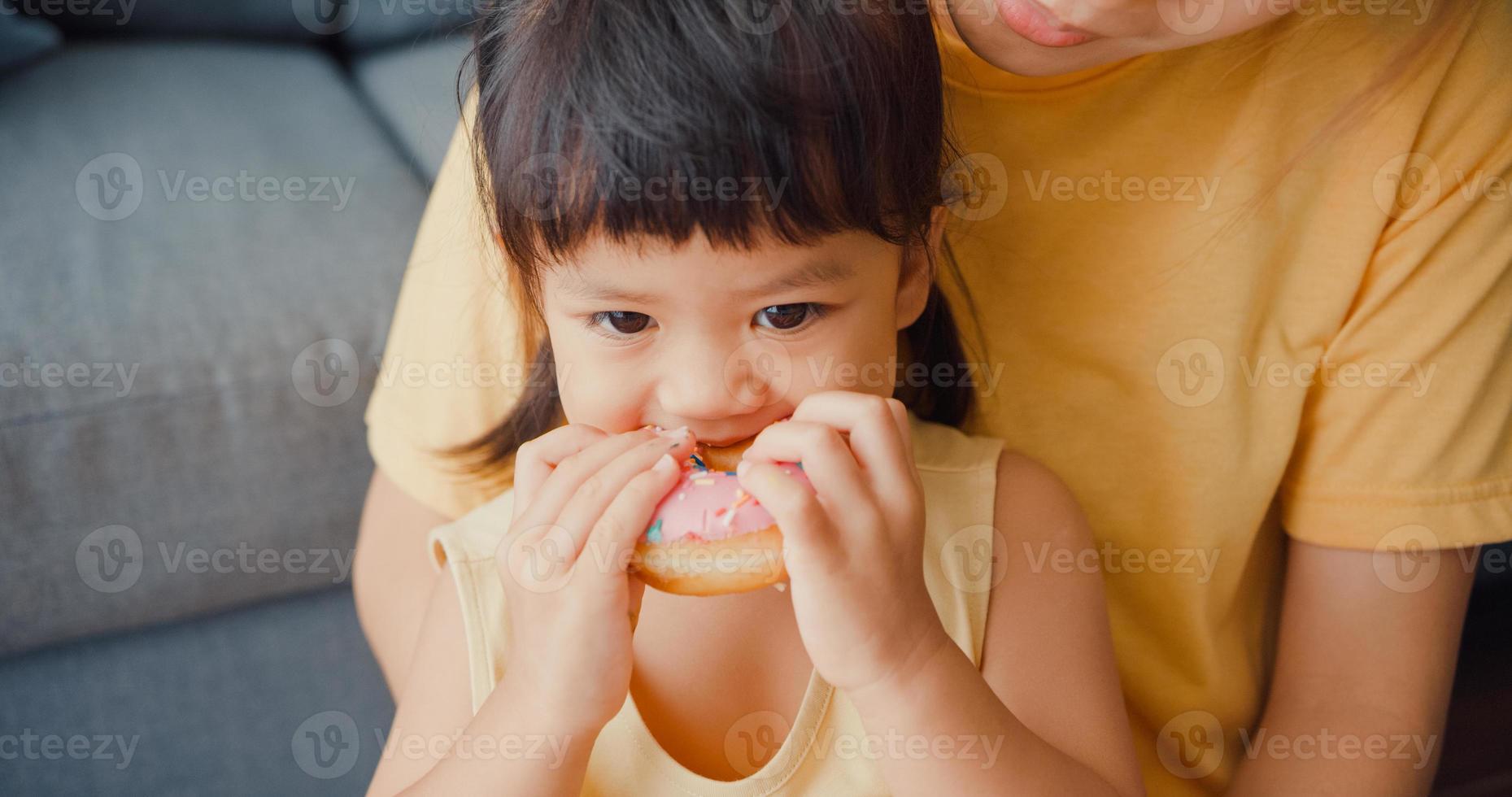 felice allegra famiglia asia mamma e bambina che mangiano ciambelle e si divertono rilassati goditi sul divano nel soggiorno di casa. passare del tempo insieme, distanza sociale, quarantena per il coronavirus. foto