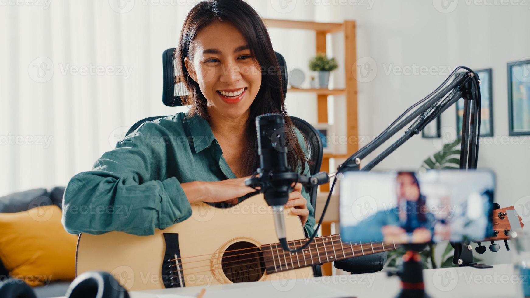 l'influencer di una ragazza asiatica adolescente suona musica per chitarra usa la registrazione del microfono con lo smartphone per il pubblico online ascolta a casa. il podcaster femminile crea podcast audio dal suo studio di casa, resta a casa il concetto. foto