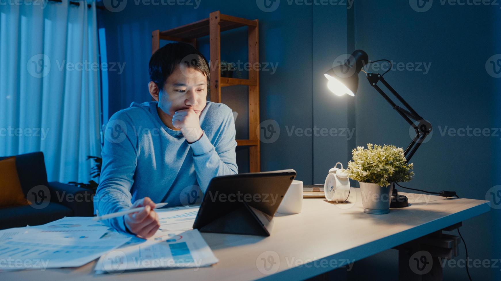 asia uomo d'affari freelance focus lavoro penna scrittura su tablet computer occupato con pieno di documenti grafici sulla scrivania in soggiorno a casa gli straordinari di notte, lavoro da casa durante il concetto di pandemia covid-19. foto