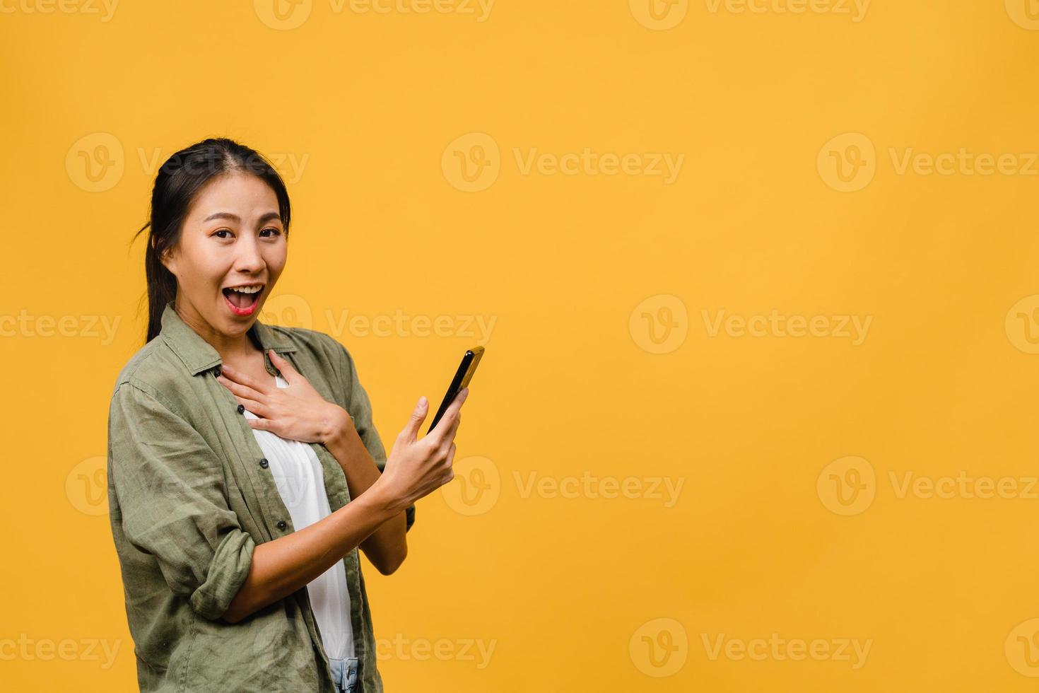 sorpresa giovane signora asiatica che utilizza il telefono cellulare con espressione positiva, sorride ampiamente, vestita con abiti casual e guardando la telecamera su sfondo giallo. felice adorabile donna felice esulta successo. foto