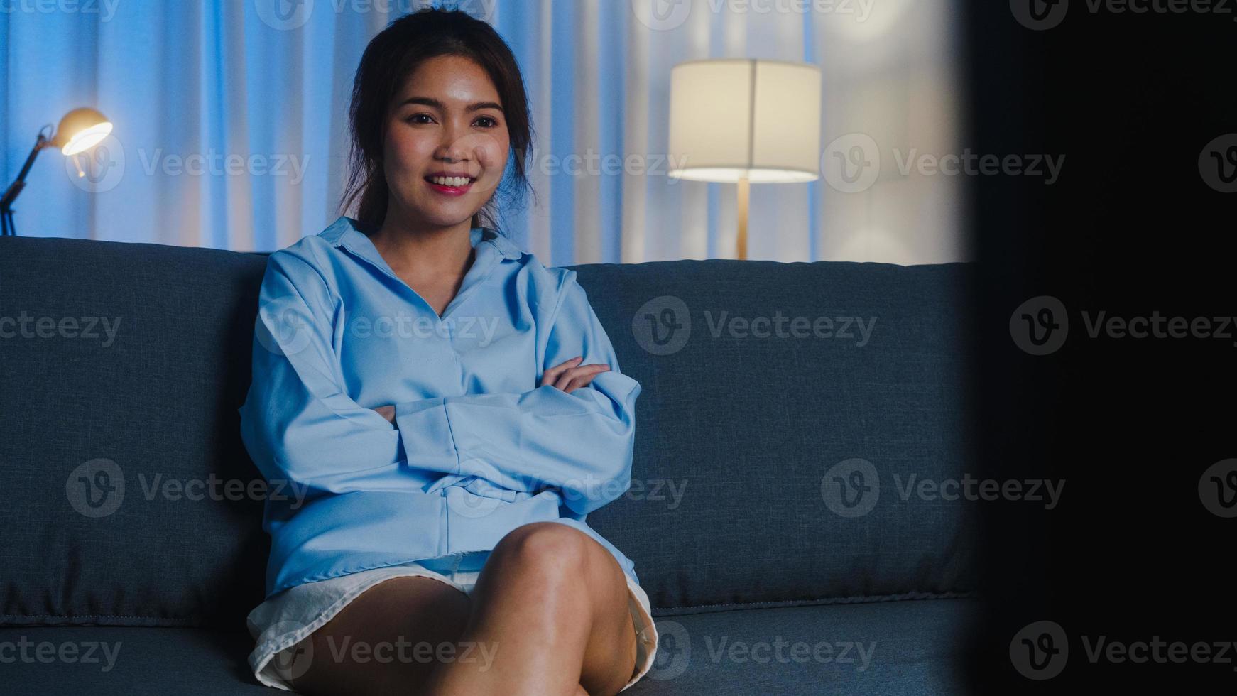 giovane donna asiatica che guarda la tv sentendosi felice e divertente ridere guardando un film di serie televisive mentre è seduta sul divano nel soggiorno di casa di notte. distanziamento sociale, quarantena per la prevenzione del coronavirus. foto