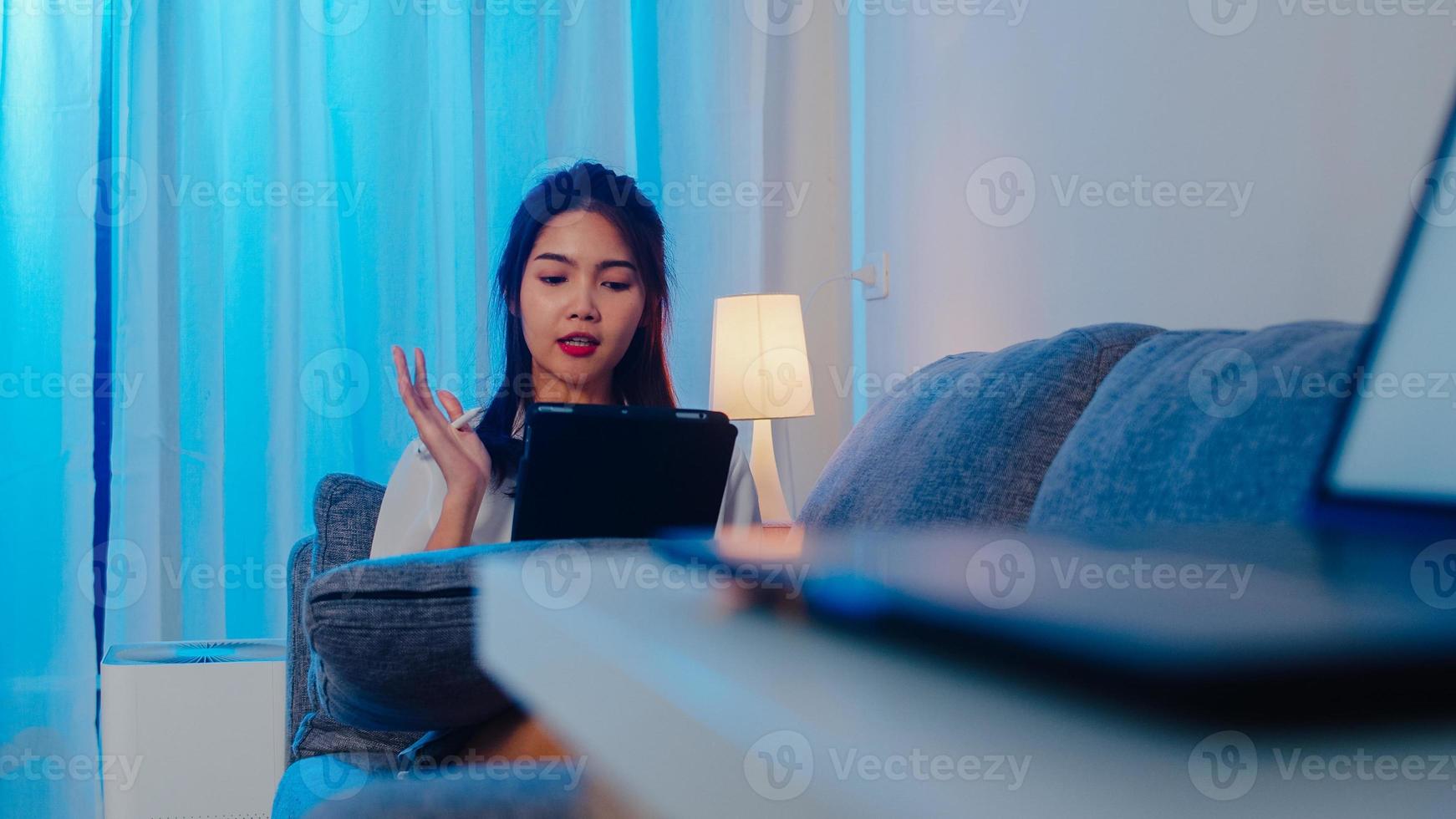 donna d'affari asiatica che utilizza tablet parla con i colleghi del piano in videochiamata mentre lavora da casa in soggiorno di notte. autoisolamento, distanziamento sociale, quarantena per la prevenzione del coronavirus. foto