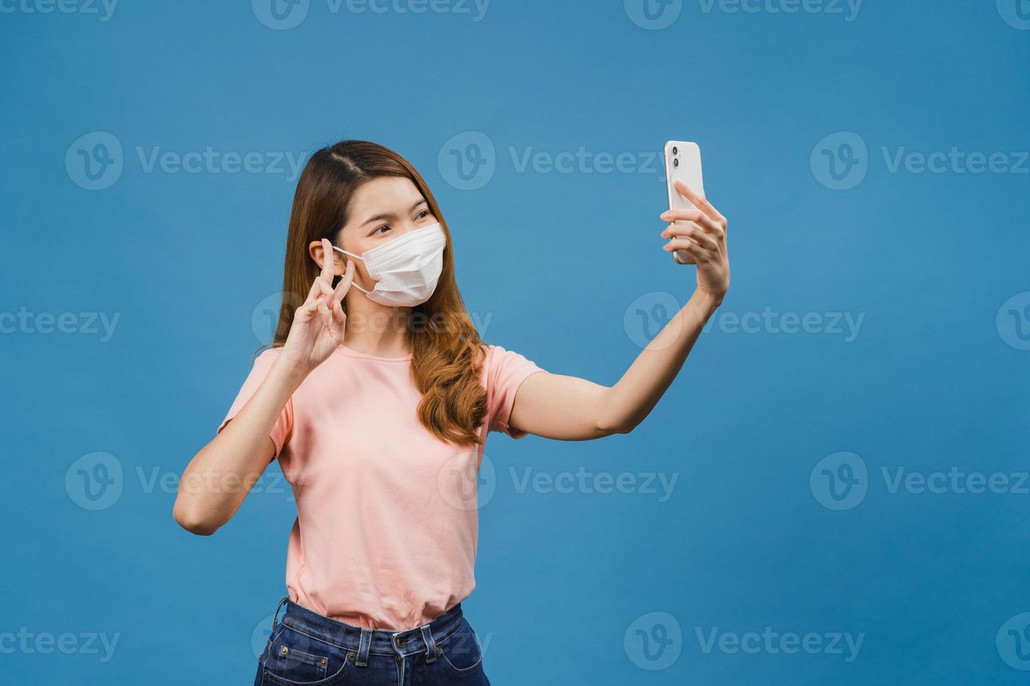 sorridente adorabile femmina asiatica che indossa maschera medica facendo selfie foto su smart phone con espressione positiva in abbigliamento casual e stand isolato su sfondo blu.