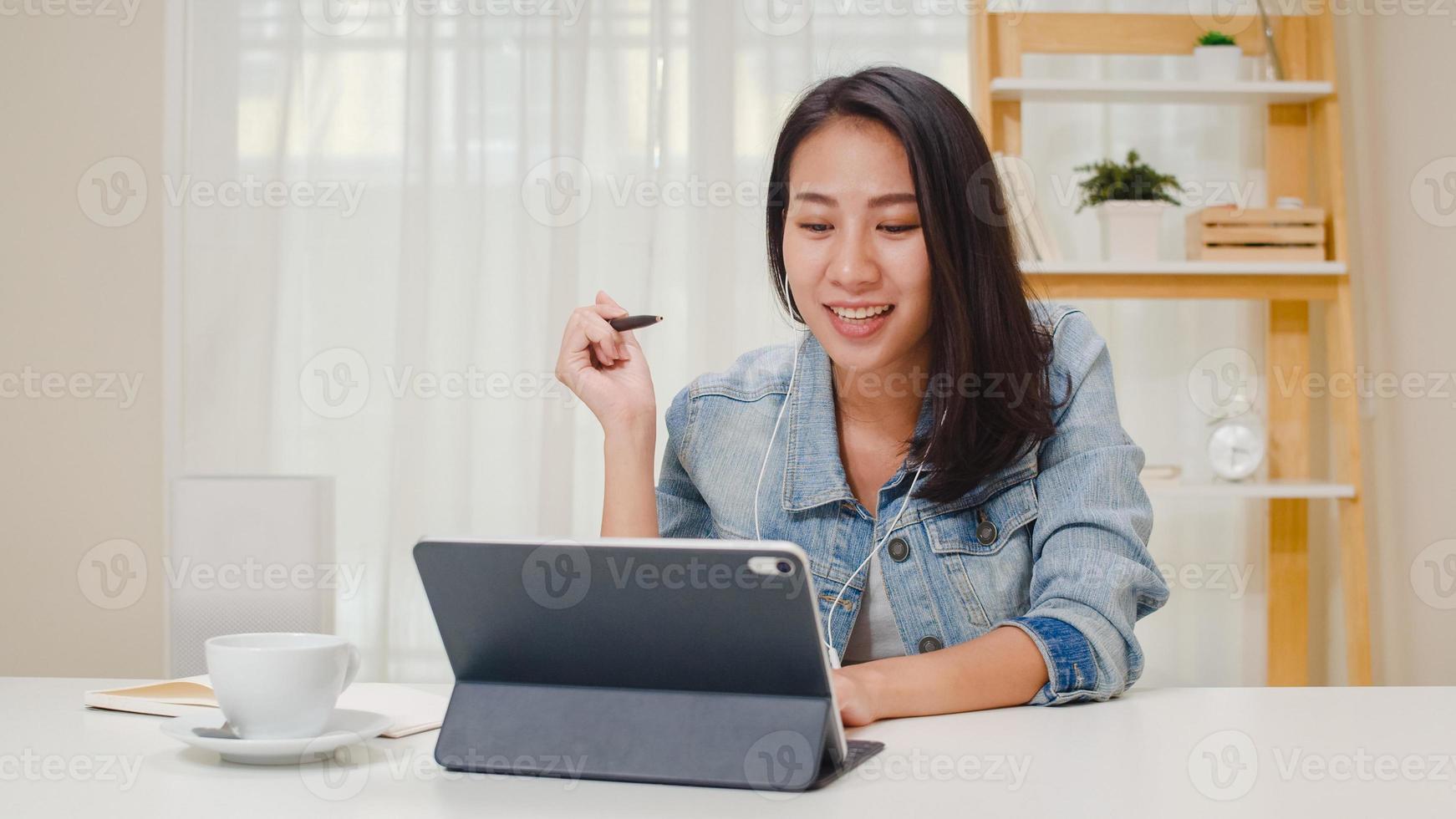 donne d'affari freelance abbigliamento casual utilizzando tablet videoconferenza chiamata di lavoro con il cliente sul posto di lavoro nel soggiorno di casa. felice giovane ragazza asiatica rilassarsi seduto sulla scrivania fare un lavoro in internet. foto
