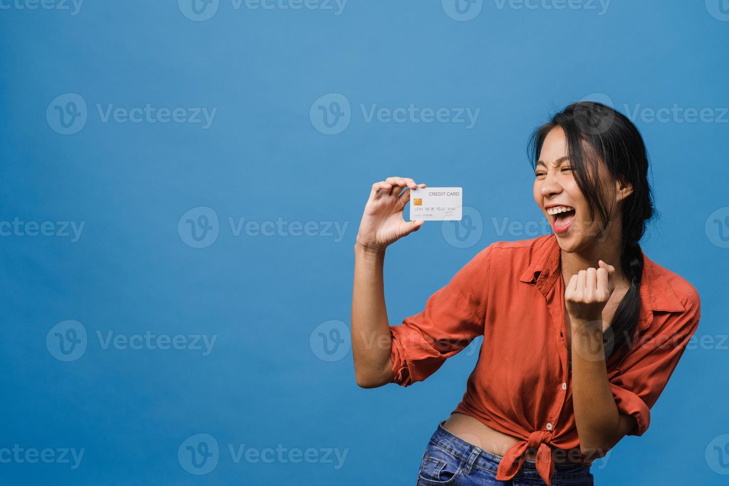 la giovane donna asiatica mostra la carta di credito con l'espressione positiva, sorride ampiamente, vestita con abiti casual sentendo felicità e stando isolata su sfondo blu. concetto di espressione facciale. foto