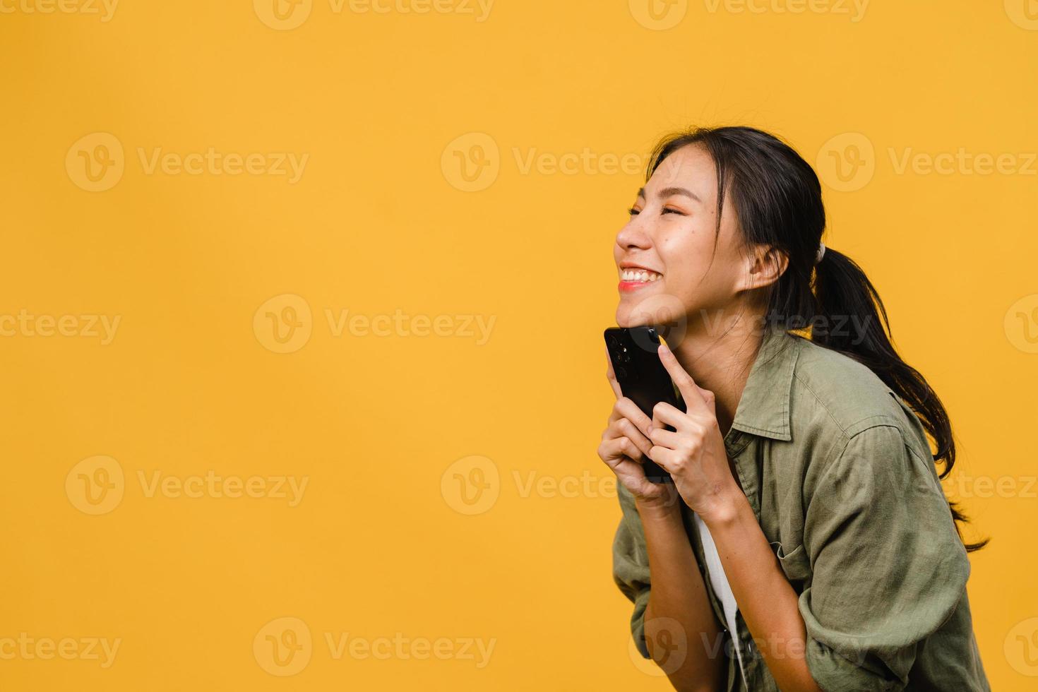 giovane donna asiatica che usa il telefono con un'espressione positiva, sorride ampiamente, vestita con abiti casual sentendosi felici e in piedi isolato su sfondo giallo. felice adorabile donna felice esulta successo. foto