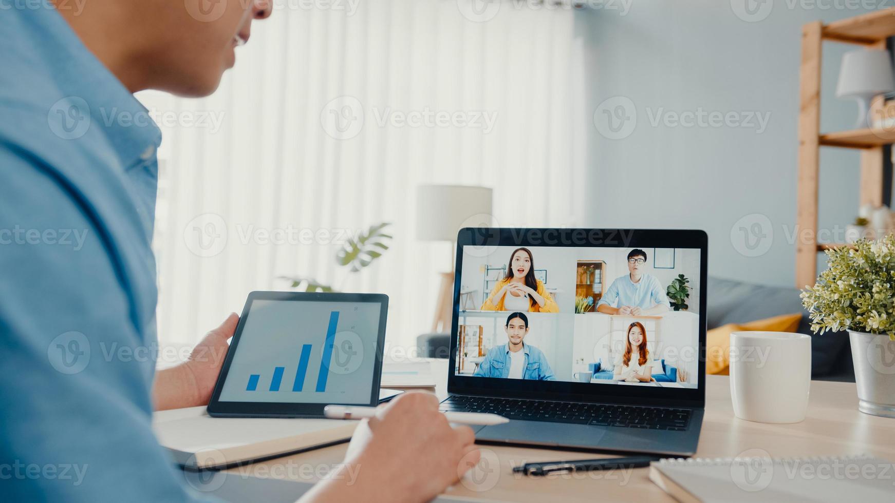 il giovane uomo d'affari asiatico che utilizza il laptop parla con i colleghi del piano in una riunione di videochiamata mentre lavora da casa in soggiorno. autoisolamento, distanziamento sociale, quarantena per la prevenzione del virus corona. foto