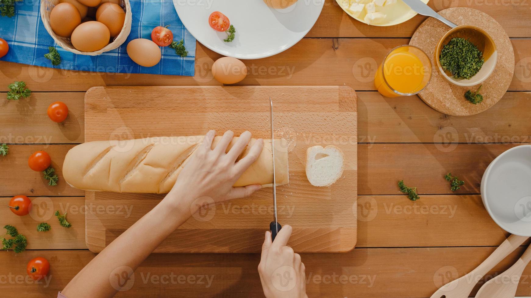 le mani della giovane donna asiatica chef tenere il coltello tagliare il pane integrale su tavola di legno sul tavolo della cucina in casa. produzione di pane fresco fatto in casa, alimentazione sana e concetto di panetteria tradizionale. colpo di vista dall'alto. foto