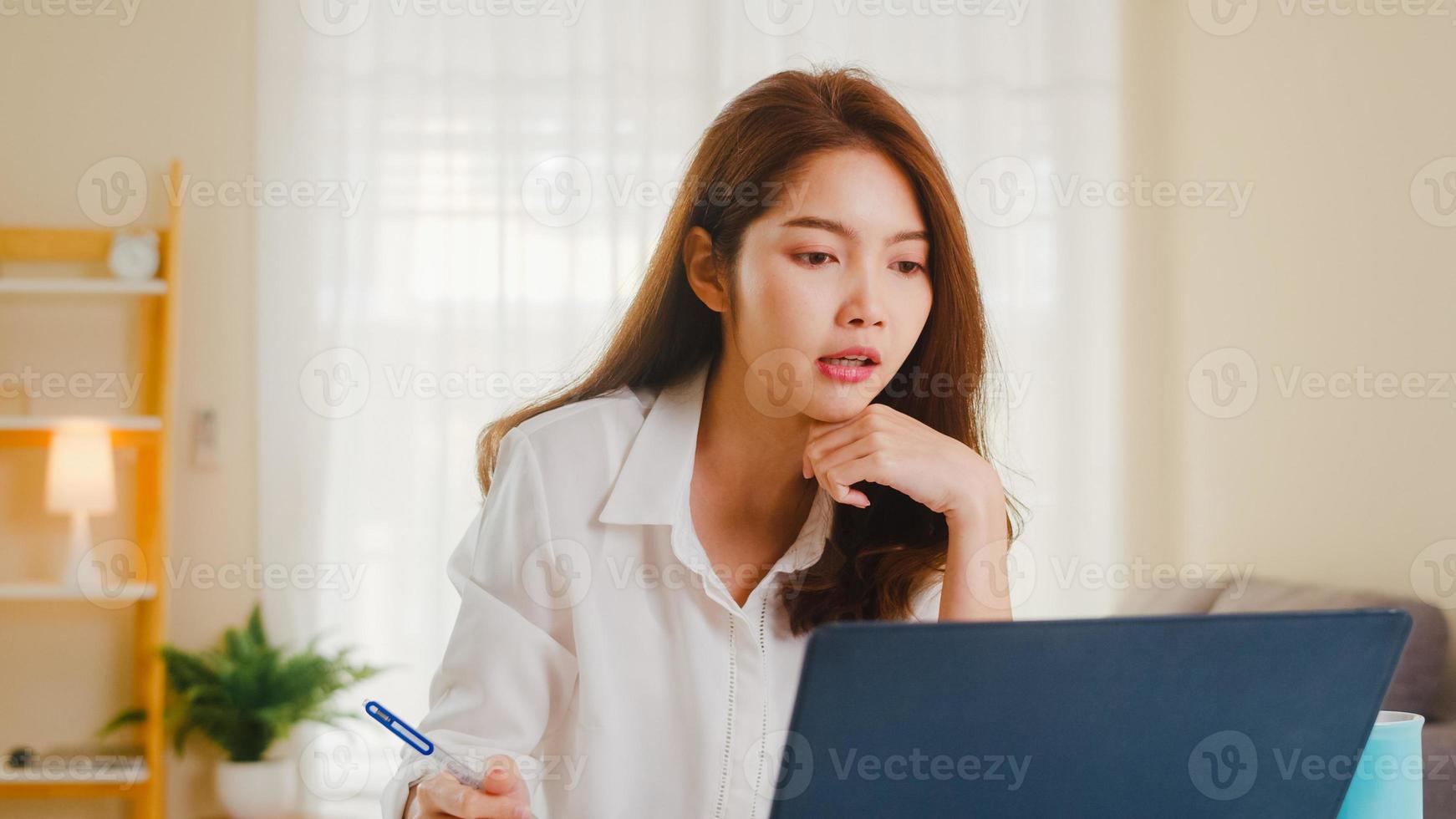 donna d'affari asiatica che utilizza laptop parla con i colleghi del piano in videochiamata mentre lavora in modo intelligente da casa in soggiorno. autoisolamento, distanziamento sociale, quarantena per la prevenzione del coronavirus. foto