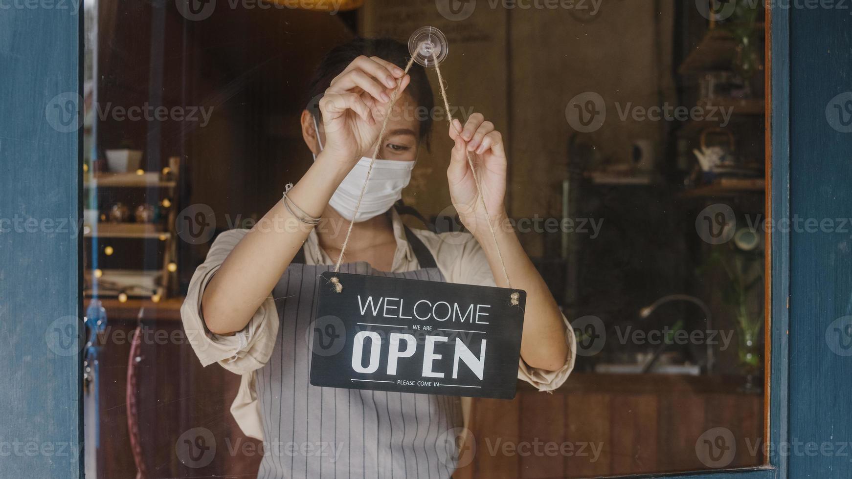 la giovane ragazza asiatica indossa la maschera per il viso girando un cartello da chiuso a aperto sulla porta che guarda fuori in attesa di clienti dopo il blocco. proprietario piccola impresa, cibo e bevande, affari riaprono concetto. foto