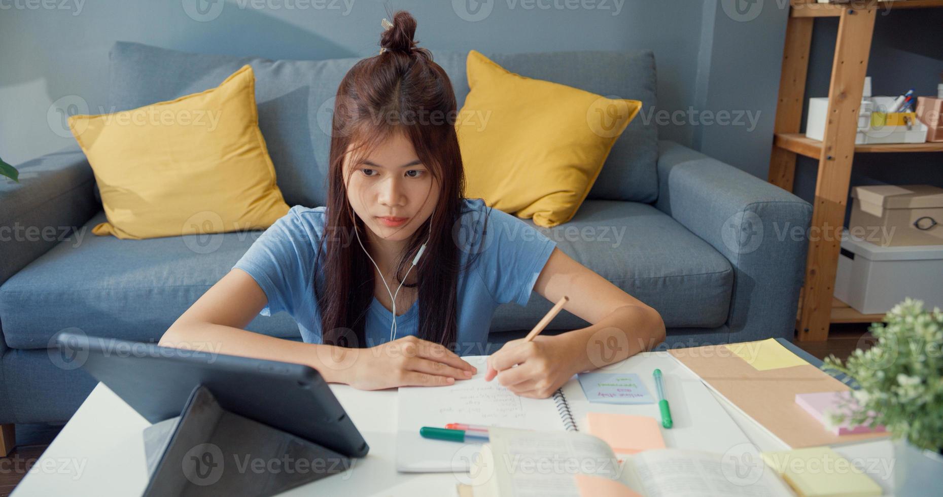 giovane ragazza asiatica adolescente con cuffie per abbigliamento casual usa tablet digitale impara taccuino di lettura online di scrittura nel soggiorno di casa. isolare l'istruzione online e-learning concetto di pandemia di coronavirus. foto