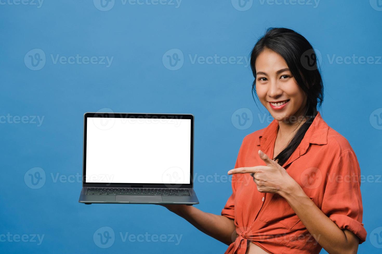 la giovane donna asiatica mostra lo schermo del laptop vuoto con un'espressione positiva, sorride ampiamente, vestita con abiti casual sentendo felicità isolata su sfondo blu. computer con schermo bianco in mano femminile. foto