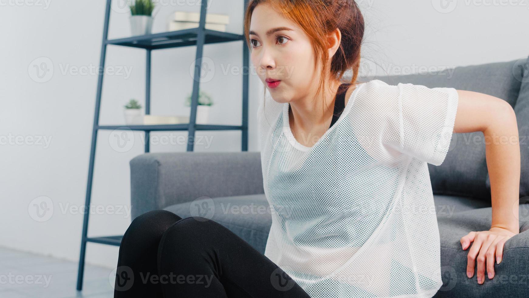 giovane donna coreana in esercizi di abbigliamento sportivo facendo allenamento facendo tuffi tricipiti appoggiata sul divano nel soggiorno di casa. distanza sociale, isolamento durante il virus. esercizi per la parte inferiore del corpo. foto