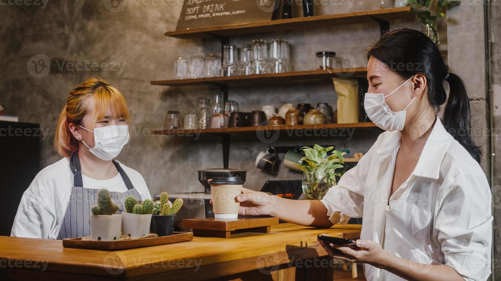 giovane barista femminile asiatico indossa una maschera facciale che serve da asporto tazza di carta da caffè caldo al consumatore al bar. proprietario piccola impresa, stile di vita nuovo normale dopo il virus corona e il concetto di distanza sociale. foto