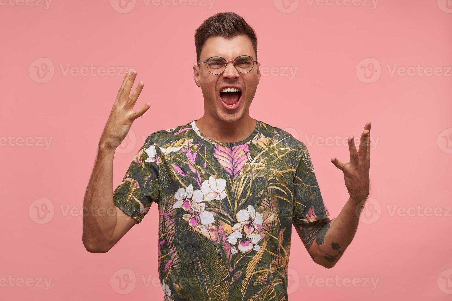 eccitato bello tipo con corto taglio di capelli in posa per telecamera con ampiamente ha aperto bocca, guardare agitato, raccolta mani, in piedi al di sopra di rosa sfondo foto