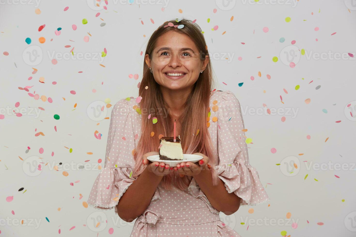 ritratto di allegro giovane bello donna con leggero Marrone lungo capelli in posa nel multicolore coriandoli con pezzo di torta nel sollevato mani, guardare verso l'alto e sorridente piacevolmente foto