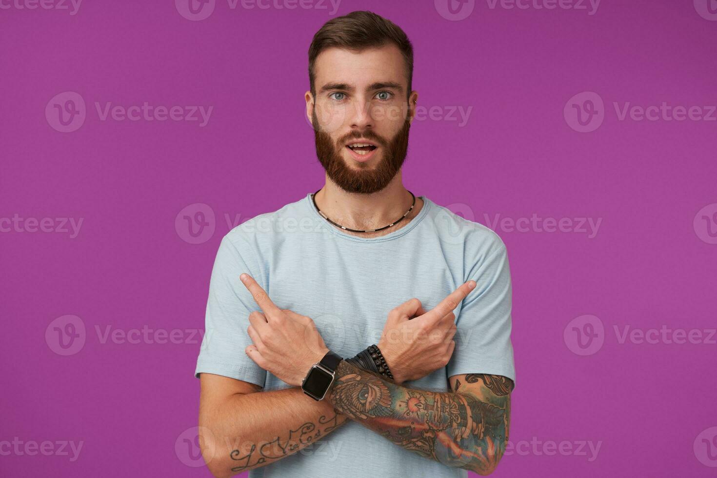 attraente giovane tatuato uomo con barba indossare blu maglietta e di moda Accessori, guardare per telecamera e mostrando nel diverso indicazioni con indice dita, isolato al di sopra di viola sfondo foto