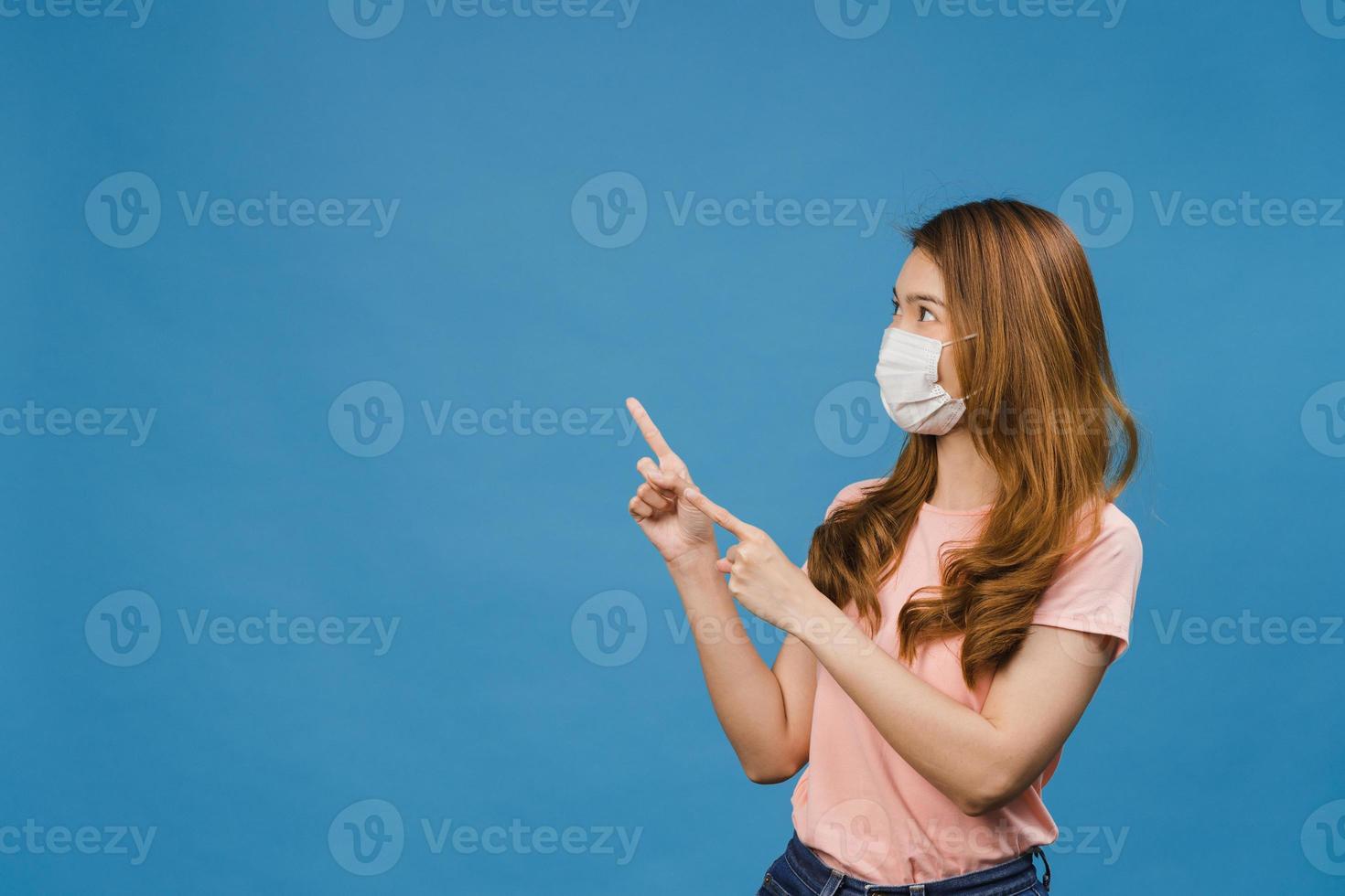 la giovane ragazza asiatica indossa una maschera medica per il viso mostra qualcosa nello spazio vuoto con un vestito casual e guarda la telecamera isolata su sfondo blu. distanziamento sociale, quarantena per il virus corona. foto