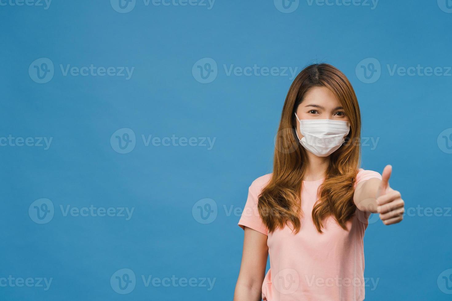 giovane ragazza asiatica che indossa una maschera medica che mostra il pollice in su con un vestito casual e guarda la telecamera isolata su sfondo blu. autoisolamento, distanziamento sociale, quarantena per il virus corona. foto