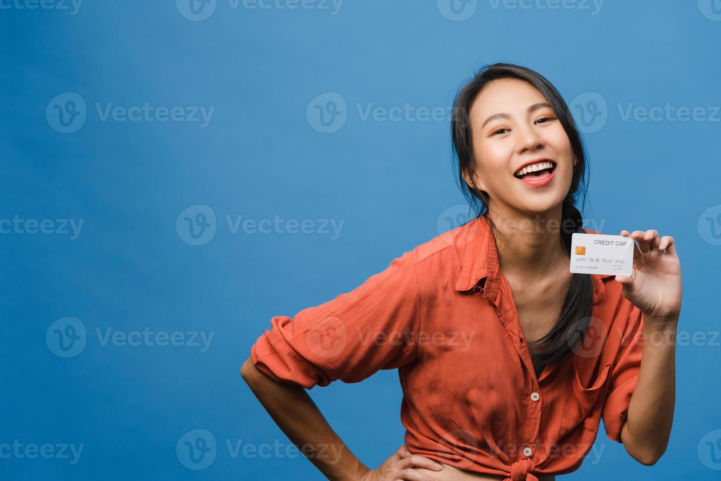 la giovane donna asiatica mostra la carta di credito con l'espressione positiva, sorride ampiamente, vestita con abiti casual sentendo felicità e stando isolata su sfondo blu. concetto di espressione facciale. foto