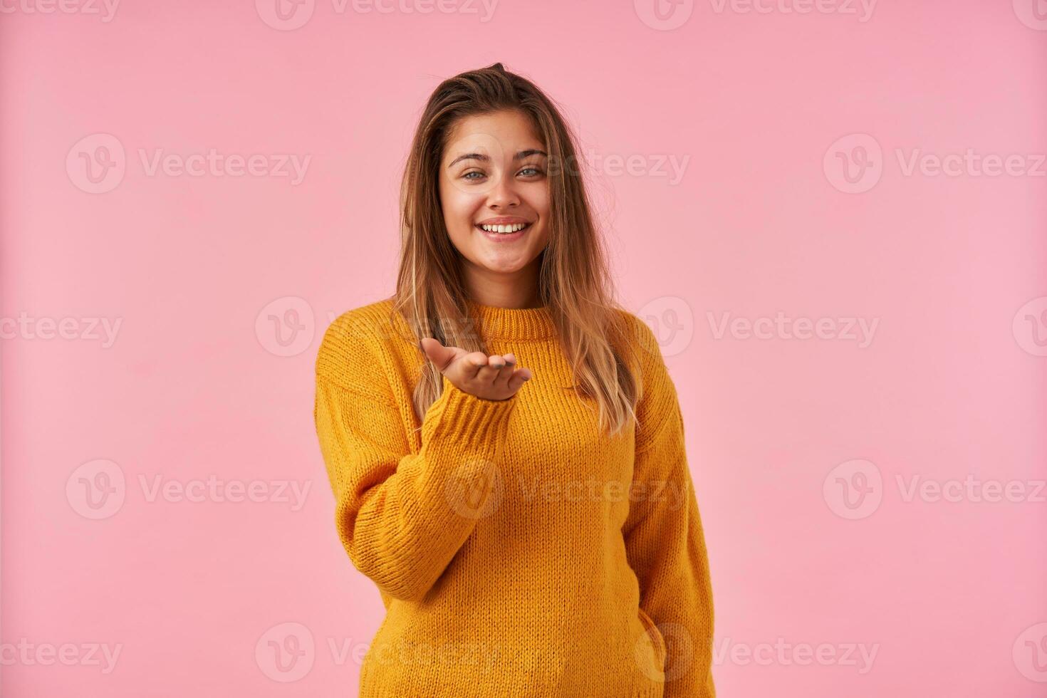 studio tiro di affascinante giovane brunetta donna guardare positivamente a telecamera con piacevole Sorridi, conservazione sua palma sollevato mentre in piedi contro rosa sfondo foto