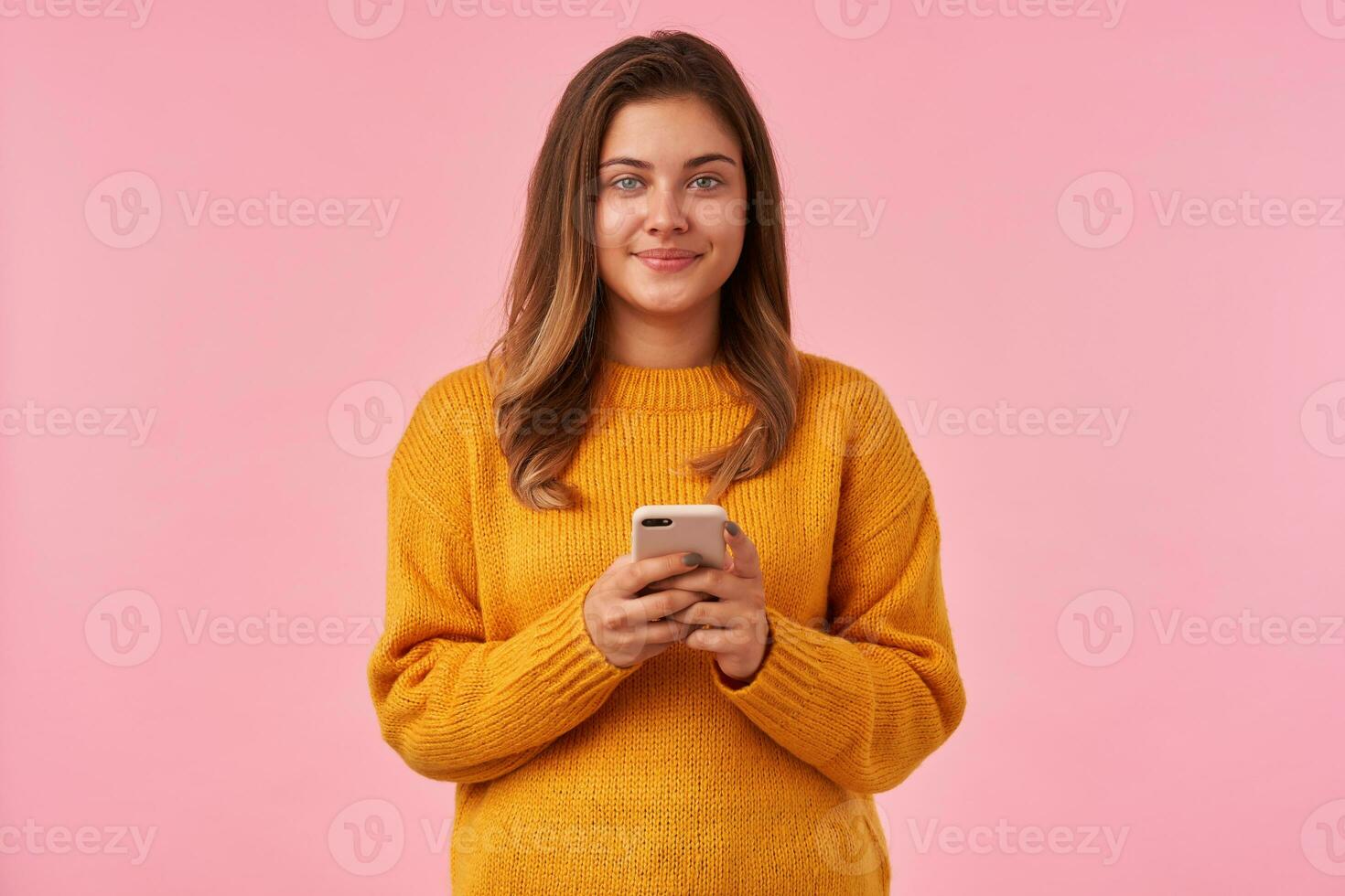 positivo giovane bello occhi verdi brunetta donna guardare a telecamera con affascinante Sorridi mentre in piedi al di sopra di rosa sfondo con mobile Telefono nel sollevato mani foto