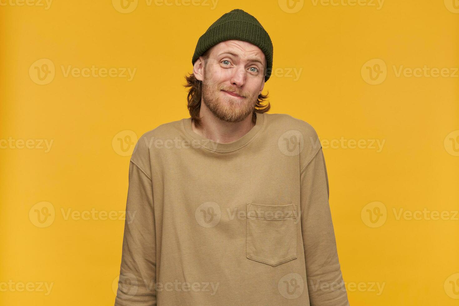 dubbioso uomo, insicuro tipo con biondo capelli, barba e baffi. indossare verde Beanie e beige maglione. alza le spalle le spalle. non fare conoscere niente. Guardando a il telecamera isolato al di sopra di giallo sfondo foto