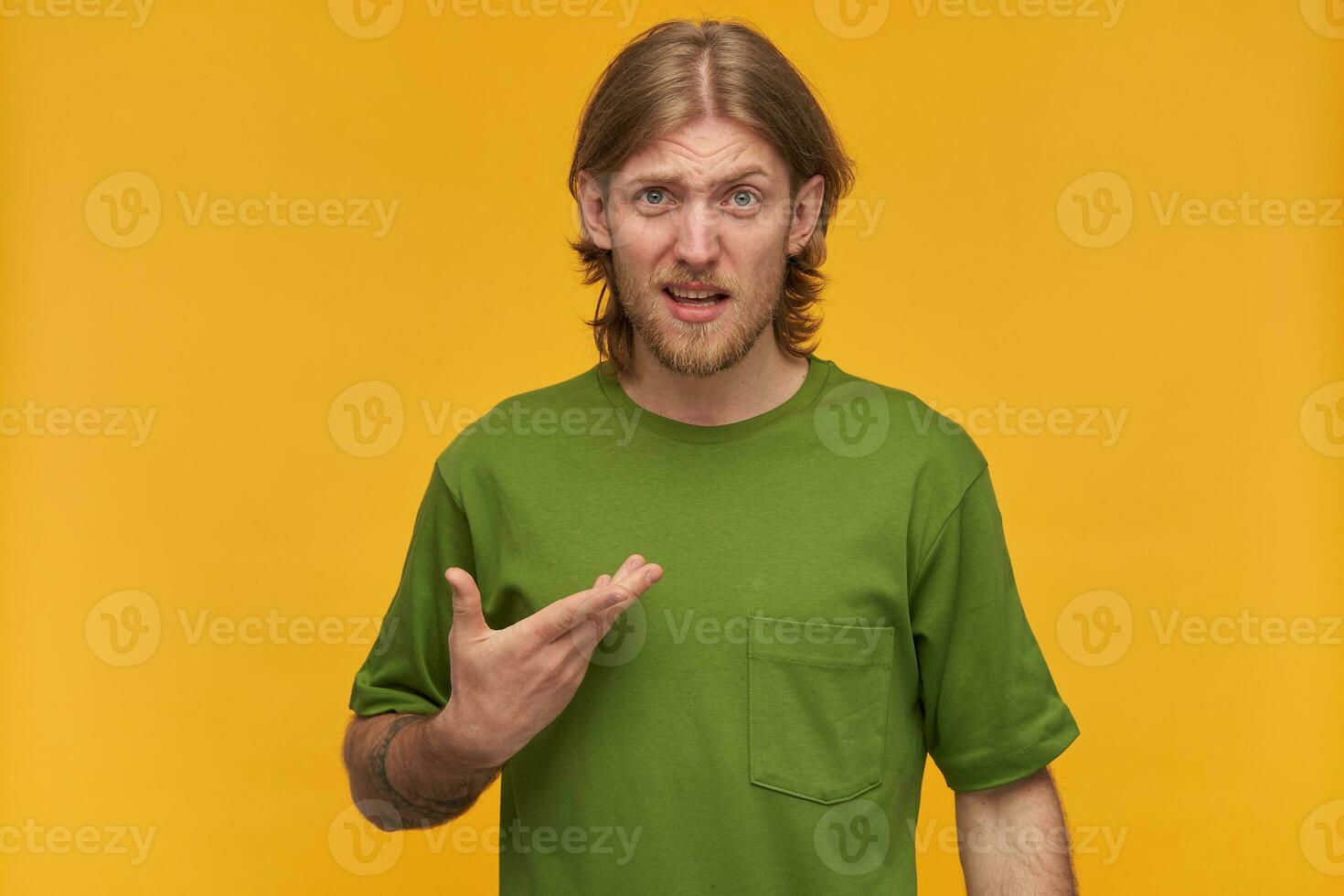 confuso uomo, infelice tipo con biondo capelli, barba e baffi. indossare verde maglietta. ha tatuaggio. puntamento a lui stesso nel smarrimento. Guardando a il telecamera isolato al di sopra di giallo sfondo foto