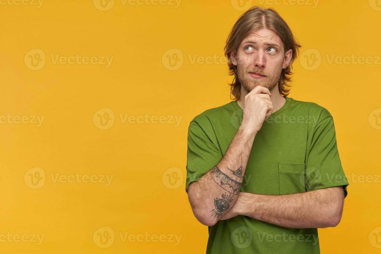 ritratto di sognare maschio con biondo acconciatura e barba. indossare verde maglietta. ha tatuaggio. toccante il suo mento. Guardando pensieroso per il sinistra a copia spazio, isolato al di sopra di giallo sfondo foto