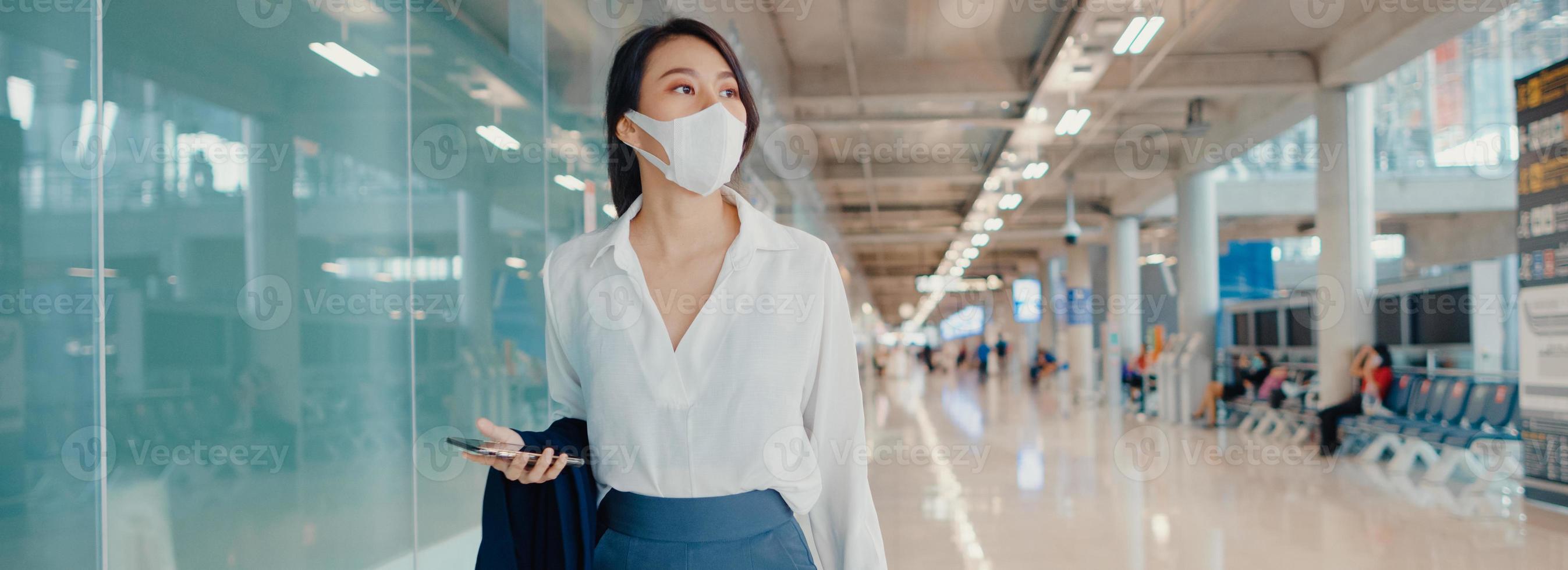 la ragazza d'affari asiatica usa lo smartphone per il check-in della carta d'imbarco a piedi con i bagagli al terminal del volo interno all'aeroporto. pandemia di covid per pendolari d'affari, sfondo panoramico banner con spazio di copia. foto