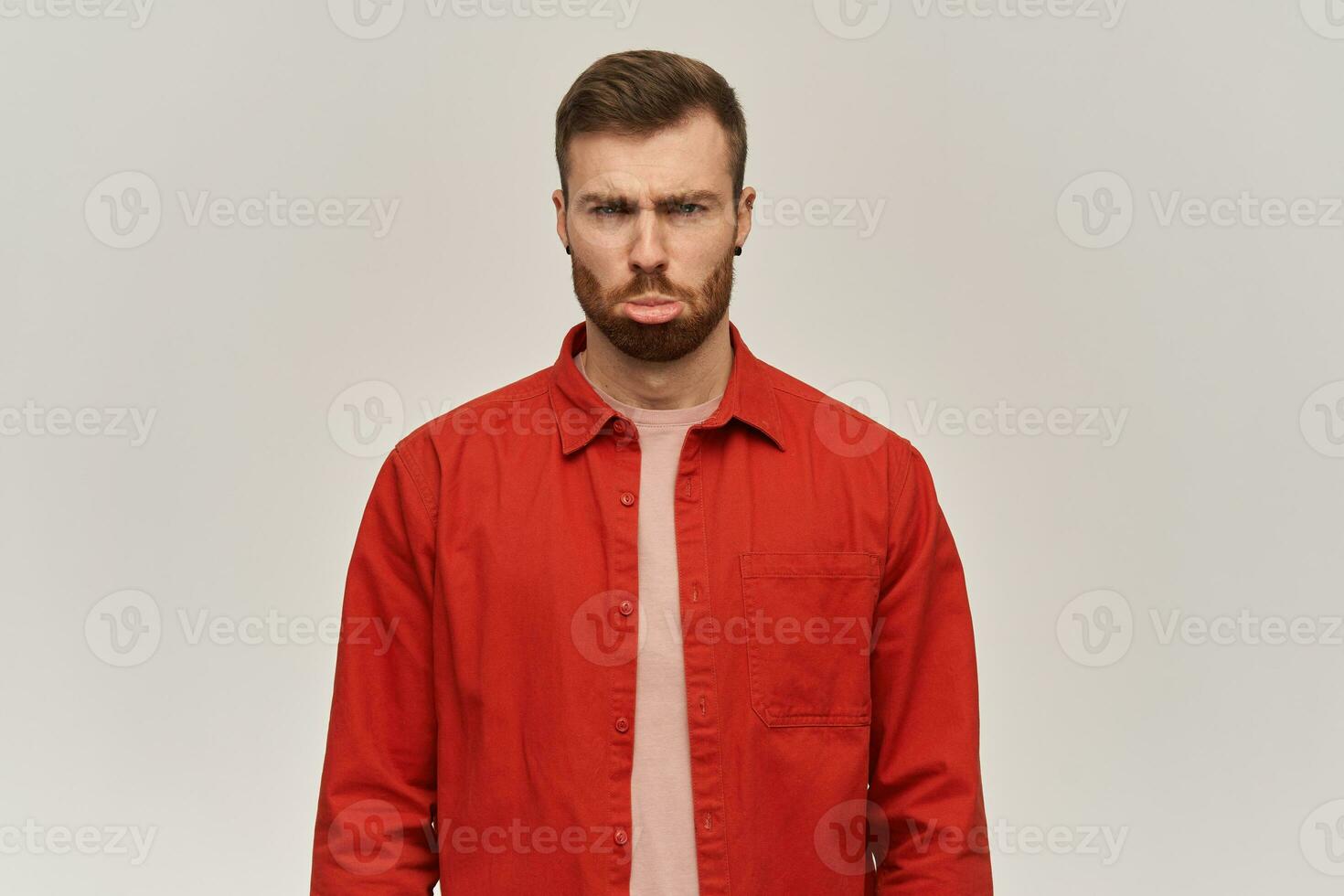 triste divertente giovane uomo nel rosso camicia con barba sembra offeso e fabbricazione divertente viso al di sopra di bianca sfondo foto